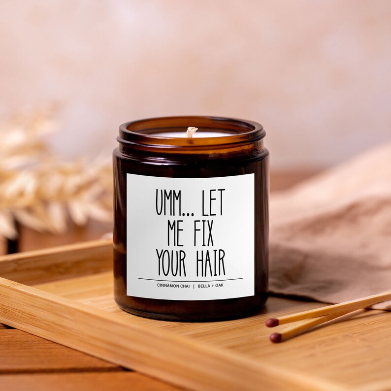 May include: A brown glass candle with a white label that reads "Umm... Let Me Fix Your Hair". The candle is on a wooden tray with a brown cloth in the background.