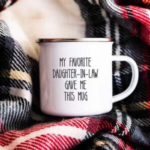 May include: White enamel mug with black text that reads "My favourite daughter-in-law gave me this mug". The mug is sitting on a red and black checked blanket.