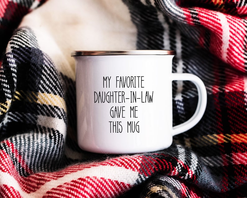 May include: White enamel mug with black text that reads "My favourite daughter-in-law gave me this mug". The mug is sitting on a red and black checked blanket.