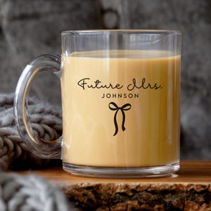 May include: A clear glass mug filled with a light brown beverage. The front of the mug features black text that reads "Future Mrs." above the name "JOHNSON" and a small black bow graphic centered below the text.