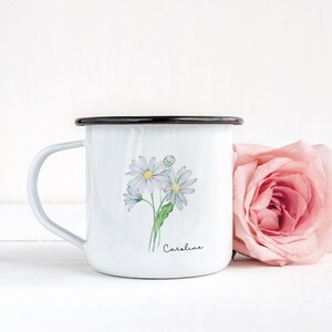 Puede incluir: Taza de esmalte blanco con borde negro y una ilustraci&oacute;n de acuarela de margaritas blancas con tallos verdes. La taza tiene el nombre "Caroline" escrito en cursiva negra en la parte inferior.