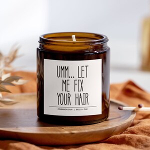 May include: A brown glass candle with a white label that reads "Umm... Let Me Fix Your Hair". The candle is on a wooden surface and has a cinnamon chai and bella + oak scent.