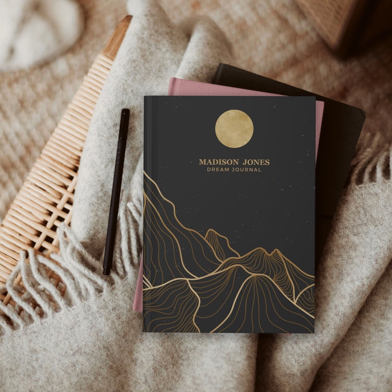 May include: A black dream journal with a gold moon and mountain design. The text on the cover reads "MADISON JONES DREAM JOURNAL".