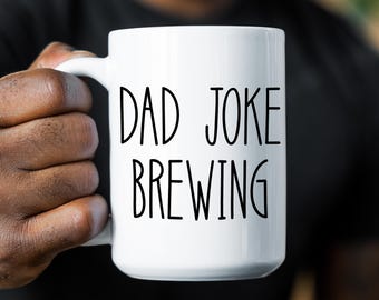May include: A white ceramic mug featuring the black text DAD JOKE BREWING printed in a simple sans-serif font. The mug has a smooth finish, a curved handle on the left side, and a cylindrical shape with a flat base, designed for holding hot beverages.