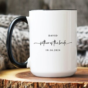 May include: A white ceramic mug with a black handle and interior rim. The front features black text that reads DAVID, followed by the script text father of the bride, and the date 10.16.2024 printed in a simple sans-serif font.