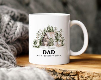 May include: A white ceramic mug featuring a watercolor illustration of a cabin nestled among pine trees. Below the cabin, the word DAD is printed in bold black capital letters, followed by the names NOAH, BETHANY, and OLIVIA in smaller text.