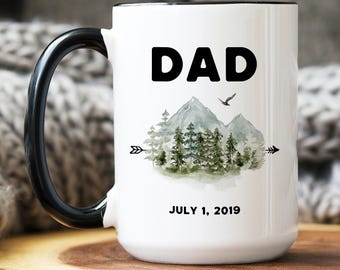 May include: A white ceramic mug with a black handle and rim. The front features the word DAD in bold black letters above a watercolor illustration of mountains, pine trees, and a bird, flanked by arrows. The text JULY 1, 2019 is printed below.