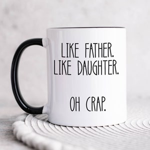 May include: White ceramic coffee mug with a black handle and black rim. The mug has the text "Like father. Like daughter. Oh crap." printed in black.