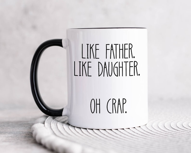 May include: White ceramic coffee mug with a black handle and black rim. The mug has the text "Like father. Like daughter. Oh crap." printed in black.