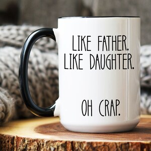 May include: White ceramic coffee mug with a black handle and black rim. The mug has the text "Like father. Like daughter. Oh crap." printed in black.