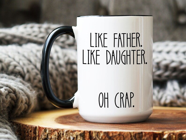 May include: White ceramic coffee mug with a black handle and black rim. The mug has the text "Like father. Like daughter. Oh crap." printed in black.