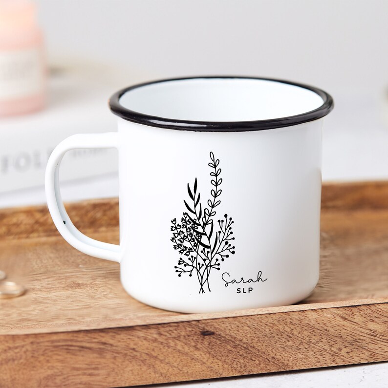 May include: White enamel mug with a black rim and handle. The mug features a black floral design and the name "Sarah" with "SLP" underneath. The mug is sitting on a wooden tray.