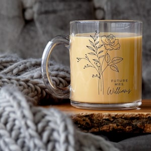 May include: A clear glass mug filled with a light tan beverage, featuring a black line art illustration of a rose and leafy stems. The text "FUTURE MRS. WILLIAMS" is printed in black lettering on the side of the mug, positioned below the floral design.