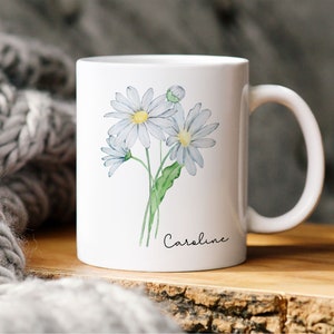 Puede incluir: Taza de cer&aacute;mica blanca con una ilustraci&oacute;n de acuarela de tres margaritas blancas y tallos verdes. La taza tiene el nombre "Caroline" escrito en cursiva negra en la parte inferior.