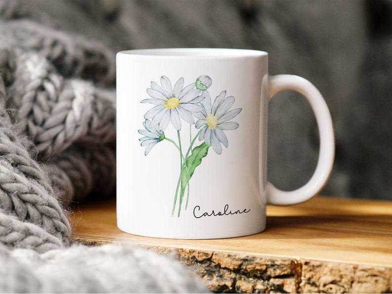 Puede incluir: Taza de cer&aacute;mica blanca con una ilustraci&oacute;n de acuarela de tres margaritas blancas y tallos verdes. La taza tiene el nombre "Caroline" escrito en cursiva negra en la parte inferior.