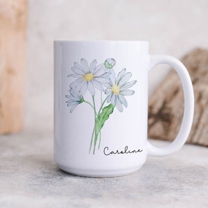 Puede incluir: Taza de cer&aacute;mica blanca con un dise&ntilde;o floral que presenta tres margaritas blancas con centros amarillos y tallos verdes. La taza tiene un asa negra y el nombre "Caroline" est&aacute; escrito en cursiva negra en la parte inferior.