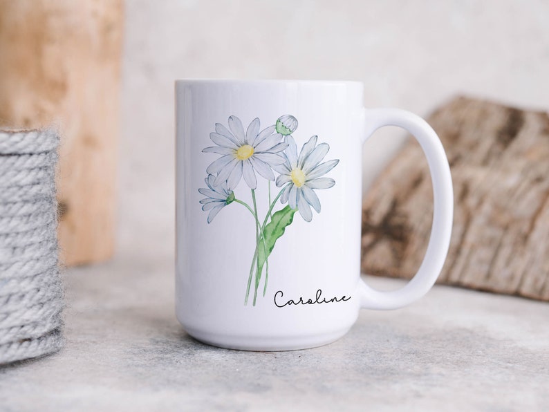 Puede incluir: Taza de cer&aacute;mica blanca con un dise&ntilde;o floral que presenta tres margaritas blancas con centros amarillos y tallos verdes. La taza tiene un asa negra y el nombre "Caroline" est&aacute; escrito en cursiva negra en la parte inferior.