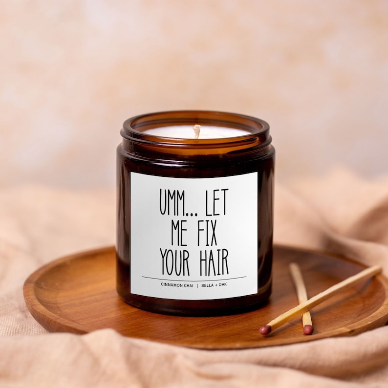 May include: A brown glass candle jar with a white label that reads "Umm... Let Me Fix Your Hair". The candle is on a wooden tray and has a white wick.