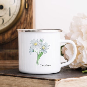 Puede incluir: Taza de esmalte blanco con borde plateado y una ilustraci&oacute;n de acuarela de margaritas blancas con tallos verdes. La taza tiene el nombre "Caroline" escrito en cursiva negra en la parte inferior.