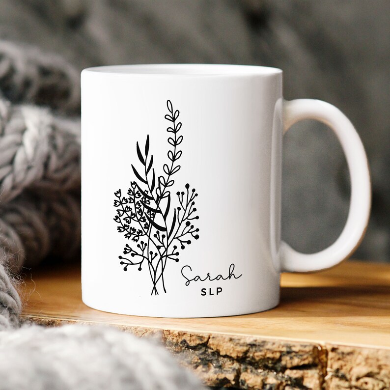 May include: White ceramic mug featuring a black floral design with the name "Sarah" and the initials "SLP" in a delicate script. The mug has a curved handle and is set against a blurred background.