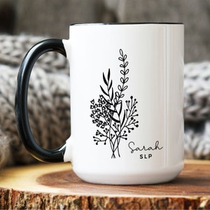 May include: White ceramic mug with a black handle and rim. The mug features a black floral design and the name "Sarah" with "SLP" underneath. The mug is sitting on a wooden surface.