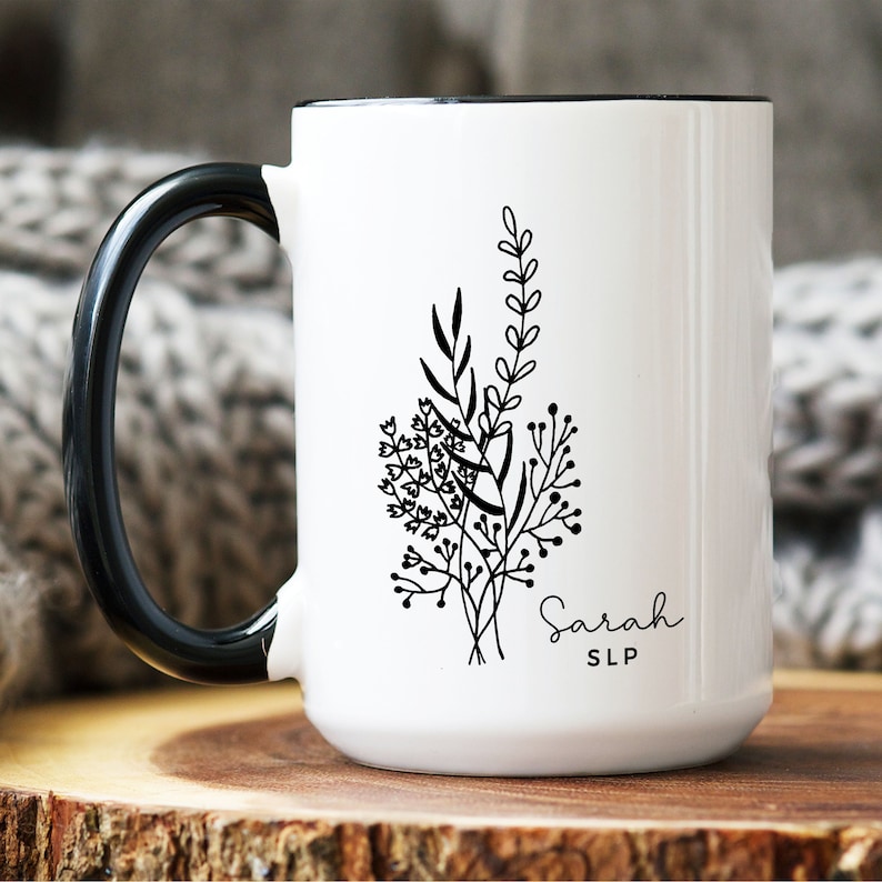 May include: White ceramic mug with a black handle and rim. The mug features a black floral design and the name "Sarah" with "SLP" underneath. The mug is sitting on a wooden surface.