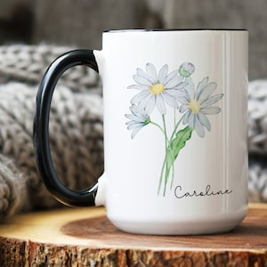 Puede incluir: Taza de cer&aacute;mica blanca con asa negra. La taza presenta una ilustraci&oacute;n de acuarela de un ramo de margaritas blancas con tallos y hojas verdes. El texto "Caroline" est&aacute; impreso en cursiva negra en la parte inferior de la taza.
