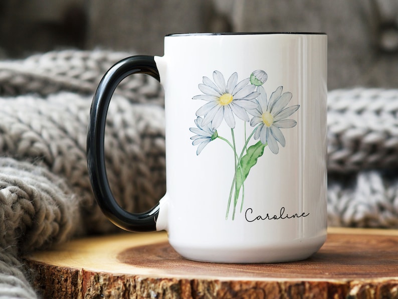 Puede incluir: Taza de cer&aacute;mica blanca con asa negra. La taza presenta una ilustraci&oacute;n de acuarela de un ramo de margaritas blancas con tallos y hojas verdes. El texto "Caroline" est&aacute; impreso en cursiva negra en la parte inferior de la taza.