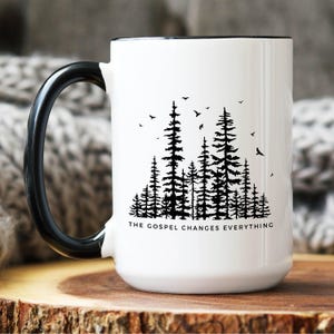 May include: White ceramic mug with a black handle and rim. The mug features a black silhouette of a forest with the text "The Gospel Changes Everything" at the bottom.