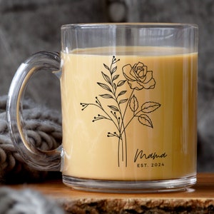 May include: A clear glass mug with a black floral design and the text "Mama Est. 2024". The mug is filled with a light brown liquid.