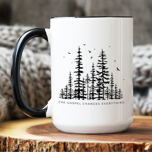 May include: A white ceramic mug with a black handle and rim. The front features a black graphic of a dense pine forest with birds flying above the trees. Below the forest, the text reads, THE GOSPEL CHANGES EVERYTHING in black capital letters.