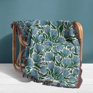 May include: A woven throw blanket with a floral pattern of blue flowers and green leaves on an off-white background. The blanket is draped over a wicker chair and has a fringed edge.