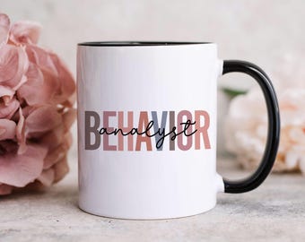 May include: A white ceramic mug with a black handle and interior rim. The front features the word BEHAVIOR in large, blocky, multi-colored letters in shades of brown, blue, and pink, with the word analyst written in black cursive script.