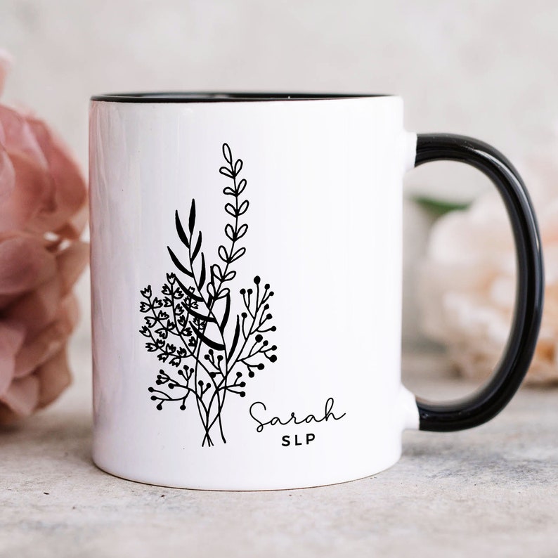 May include: White ceramic mug with a black interior and handle. The mug features a black and white floral design with the name "Sarah" and the letters "SLP" in a delicate script. The design is a simple, elegant botanical illustration.