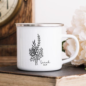 May include: White enamel mug with a silver rim and handle. The mug features a black floral design with the name "Sarah" and the initials "SLP" in a delicate script. The mug is sitting on a stack of books.