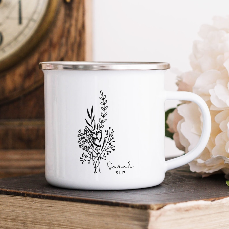 May include: White enamel mug with a silver rim and handle. The mug features a black floral design with the name "Sarah" and the initials "SLP" in a delicate script. The mug is sitting on a stack of books.