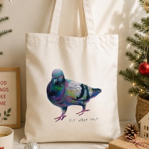 May include: A cream-coloured canvas tote bag featuring a colourful, painterly illustration of a pigeon. The bird displays iridescent shades of blue, purple, and green. Below the pigeon, the text "FLY VIBES ONLY." is printed in a simple black font.