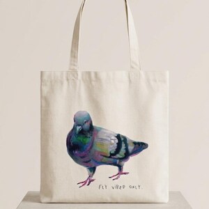 May include: A cream-coloured canvas tote bag featuring a colourful, painterly illustration of a pigeon standing in profile. Below the bird, the text "FLY VIBES ONLY." is printed in a simple, dark, handwritten-style font. The bag has two handles.