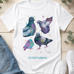 Może przedstawiać: Biały t-shirt z kr&oacute;tkim rękawem z kolorową ilustracją czterech gołębi otoczonych małymi, wielobarwnymi gwiazdkami. Napis "be kind to pigeons." jest nadrukowany w kolorze morskim pod ptakami, a "VRINDA 04.26" znajduje się w prawym dolnym rogu.