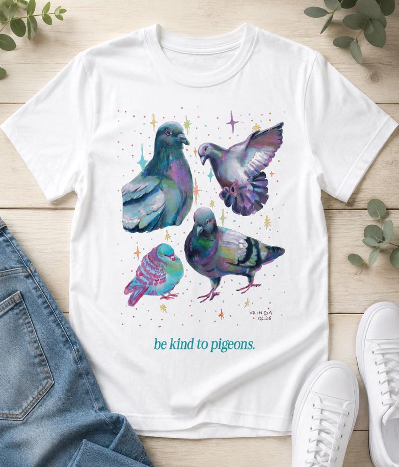 Może przedstawiać: Biały t-shirt z kr&oacute;tkim rękawem z kolorową ilustracją czterech gołębi otoczonych małymi, wielobarwnymi gwiazdkami. Napis "be kind to pigeons." jest nadrukowany w kolorze morskim pod ptakami, a "VRINDA 04.26" znajduje się w prawym dolnym rogu.