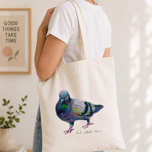 Puede incluir: Un bolso de tela color crema con una colorida ilustraci&oacute;n pict&oacute;rica de una paloma de perfil. Debajo del ave, el texto "FLY VIBED ONLY." est&aacute; impreso en la parte frontal del bolso con una tipograf&iacute;a sencilla, oscura y de estilo manuscrito.