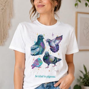 Pode incluir: Uma t-shirt branca de manga curta com um gráfico colorido de quatro pombos em várias poses, rodeados por pequenas estrelas multicoloridas. O texto "be kind to pigeons." está impresso a verde por baixo das aves, com "VRINDA 06.26".