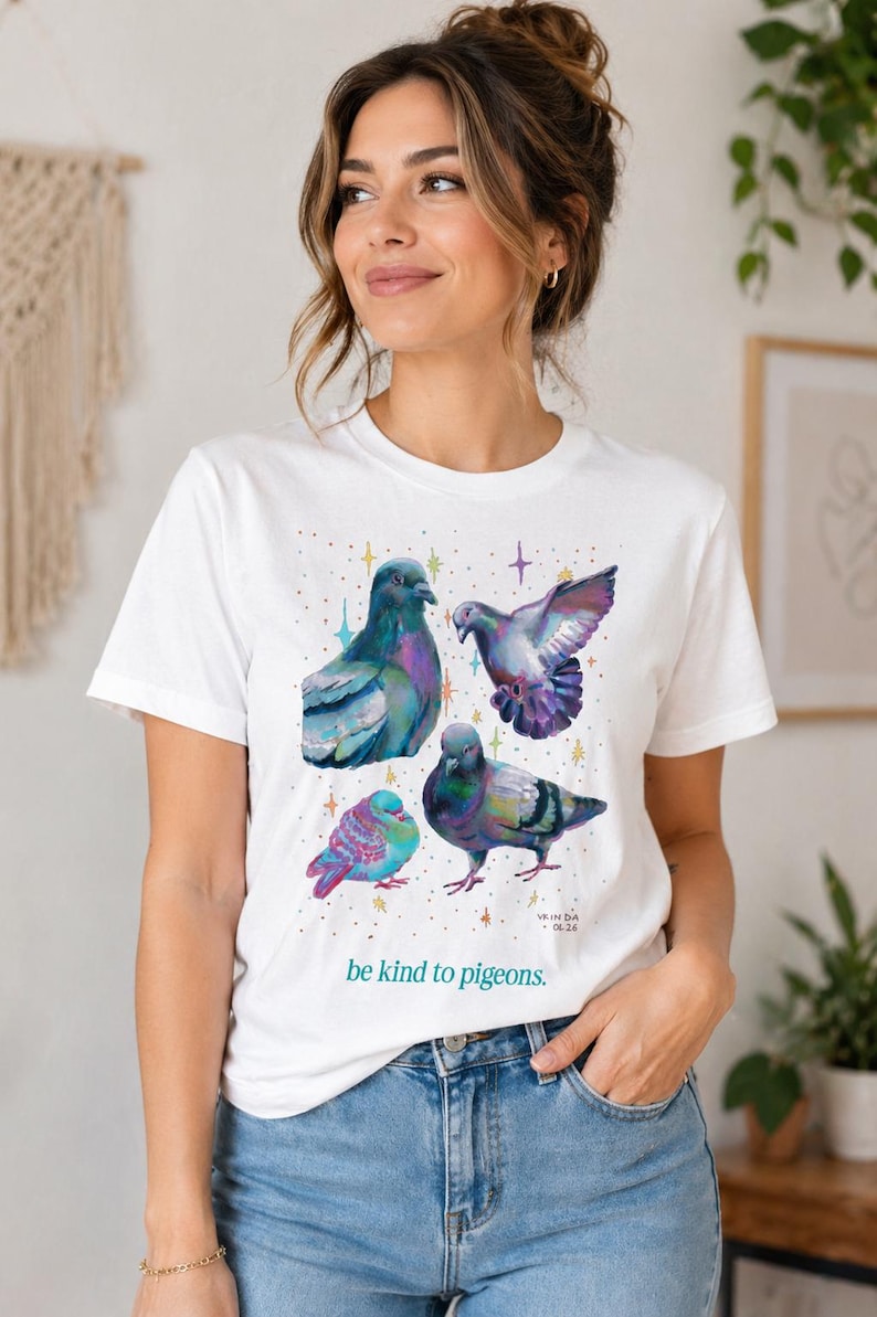 Może przedstawiać: Biały t-shirt z kr&oacute;tkim rękawem z kolorową grafiką przedstawiającą cztery gołębie w r&oacute;żnych pozach, otoczone małymi wielokolorowymi gwiazdkami. Napis "be kind to pigeons." jest wydrukowany na zielono pod ptakami, obok "VRINDA 06.26".