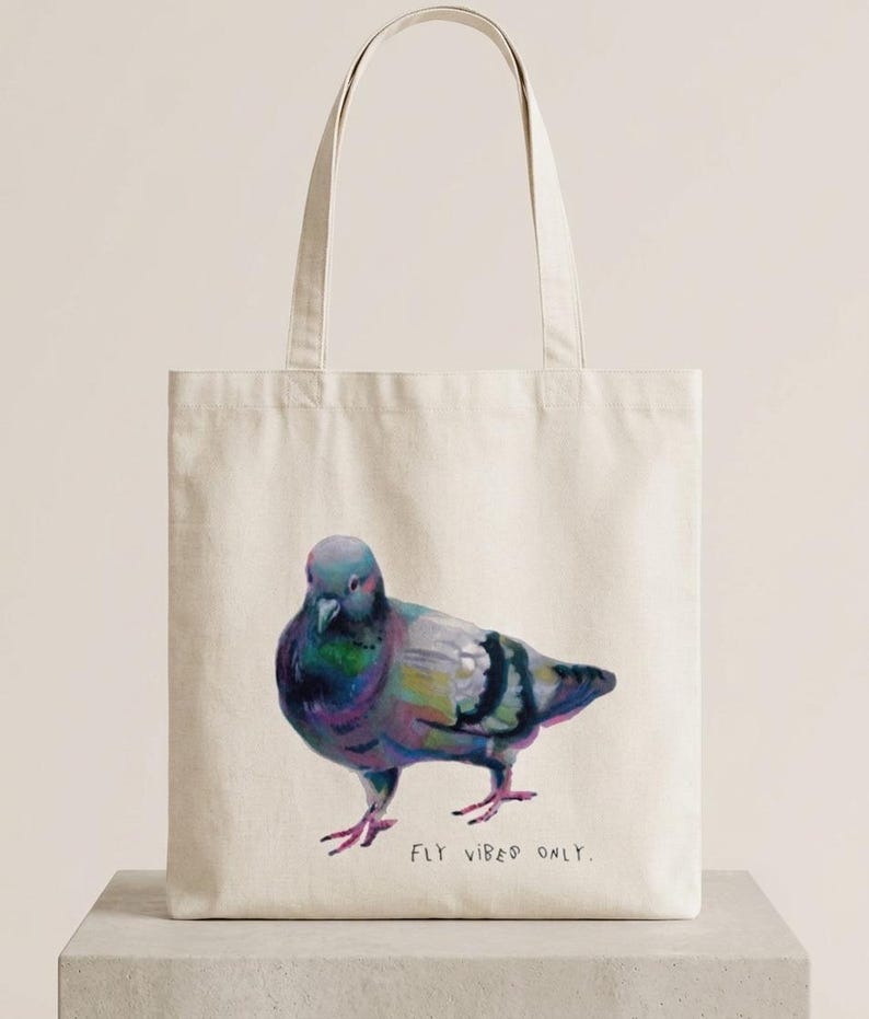 May include: A cream-coloured canvas tote bag featuring a colourful, painterly illustration of a pigeon standing in profile. Below the bird, the text "FLY VIBES ONLY." is printed in a simple, dark, handwritten-style font. The bag has two handles.