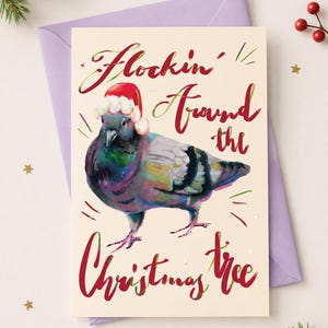 Puede incluir: Una tarjeta de felicitación con una ilustración en acuarela de una paloma con un gorro de Papá Noel rojo y blanco. El texto "Flockin' Around the Christmas tree" aparece en letra cursiva roja. La tarjeta está sobre un sobre morado claro.