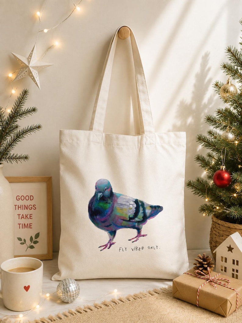 May include: A cream-coloured canvas tote bag featuring a colourful, painterly illustration of a pigeon. The bird displays iridescent shades of blue, purple, and green. Below the pigeon, the text "FLY VIBES ONLY." is printed in a simple black font.