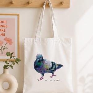 Puede incluir: Una bolsa de tela blanca con una colorida ilustraci&oacute;n pict&oacute;rica de una paloma con plumas iridiscentes de color morado, azul y verde. Debajo del ave, el texto "FLY VIBES ONLY." est&aacute; impreso en una fuente sencilla, oscura y manuscrita.