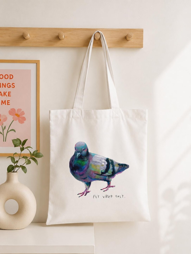 Puede incluir: Una bolsa de tela blanca con una colorida ilustraci&oacute;n pict&oacute;rica de una paloma con plumas iridiscentes de color morado, azul y verde. Debajo del ave, el texto "FLY VIBES ONLY." est&aacute; impreso en una fuente sencilla, oscura y manuscrita.
