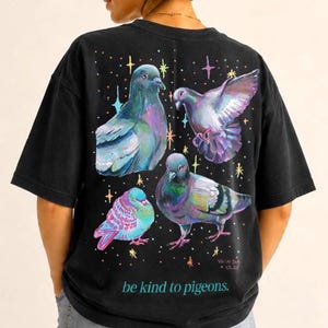 Pode incluir: Uma t-shirt preta de manga curta com um gráfico colorido de quatro pombos em várias poses, rodeados por pequenas estrelas multicoloridas. O texto "be kind to pigeons." está impresso em azul-petróleo na parte inferior do gráfico.