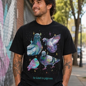 Może przedstawiać: Czarny t-shirt z kr&oacute;tkim rękawem z nadrukiem graficznym przedstawiającym cztery gołębie w opalizujących odcieniach fioletu, zieleni i błękitu, otoczone małymi kolorowymi gwiazdkami. Napis "be kind to pigeons." jest nadrukowany w kolorze morskim.